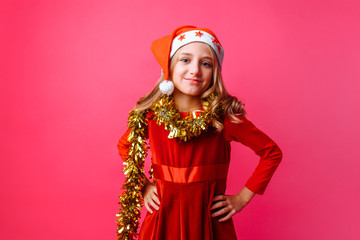 Cute teen in Santa hat and with tinsel on neck smiling on red background and in red dress. The Concept Of Christmas