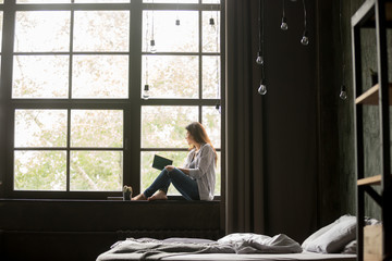 Thoughtful young brunette woman in casual clothes sitting on window sill barefoot with a book in...