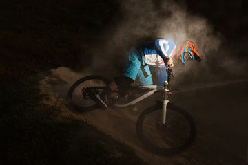 A cyclist on a mountain bike with dusty aggressive turns. Downhill riding at dark night. Bicyclist on a bicycle.
