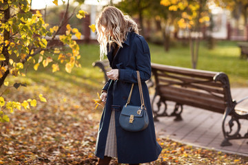 Beautiful fashionable woman walks through the autumn park in a blue coat with a bag in her hands.