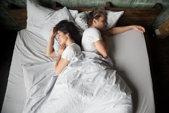 Young girl and guy, couple sleeping under blanket with their backs to each other in bed in bedroom at home, top view. Early morning, lazy Sunday weekend or day napping. People relaxing resting at home