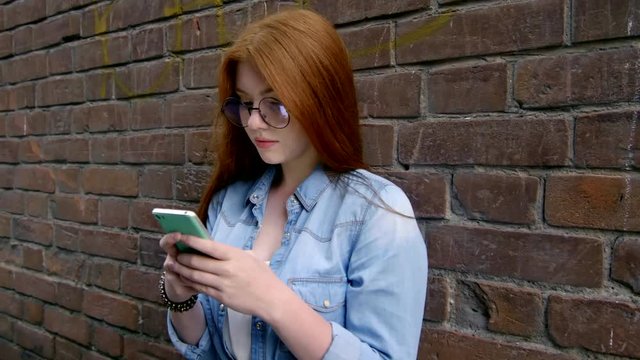 Pretty redhead teenage girl of Caucasian ethnicity standing outdoors by brick wall and typing a message on modern smartphone