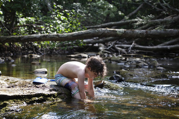Fototapeta premium Boy sitting on rock in forest river with hands in water