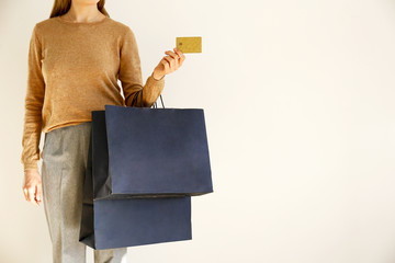 Young slim woman in classic fashionable outfit, brown sweater and loose grey pants went shopping, holding paper bags and vip golden credit / debit card. White background, copy space, close up.
