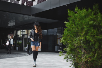 Beautiful, sexy and fashionable brunette model girl with bright makeup, in the jeans shorts, in a leather jacket and in a hat, walking outdoors with a stylish black backpack in her hand