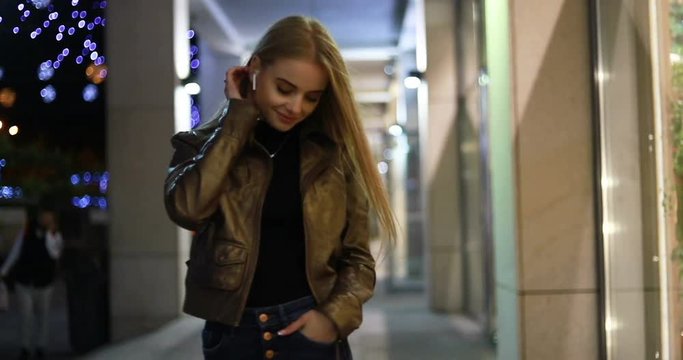 Beautiful woman looking to a clothes on a city showcase, she want it, slow motion. Young teenager girl looking at shop window at shopping mall. wireless earphones. Low light