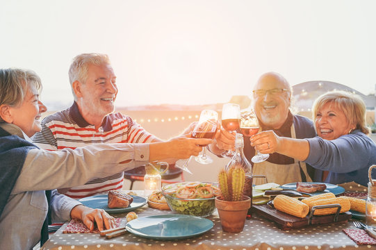 Happy senior friends having fun cheering with red wine at barbecue in terrace  outdoor - Mature people making dinner toasting glasses and laughing together - Friendship and elderly lifestyle concept