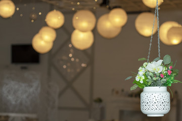 White, suspended pot with flowers against the background of round electric small lamps.