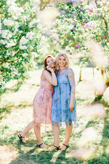 Adorable happy cheerful fabulous twin sisters in different beautiful summer dresses posing outdoor.  Similar cute female models in lilac colorful bushes with bloming flowers in park portrait.  Family.