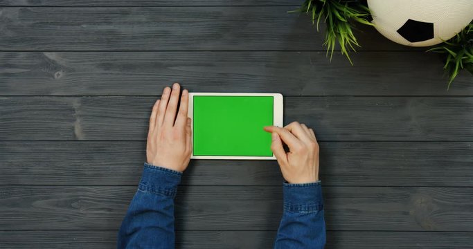Top view on the white horizontal tablet device with green screen in hands of man who scrolling and taping on the dark wooden table with football ball. Chroma key.