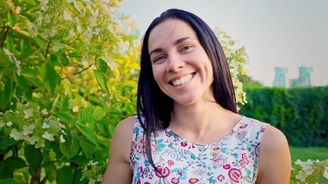 Portrait of a smiling brunette with dimpled cheeks against a green park. Beautiful young woman stands in park and looks at camera. Pretty girl in summer dress with floral print