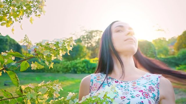 Portrait of a smiling brunette with dimpled cheeks against a green park. Beautiful young woman stands in park and looks at camera. Pretty girl in summer dress with floral print