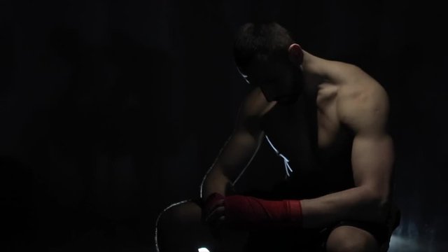 An upset muscular male boxer sitting with his head down against dark background. A topless strong sportsman