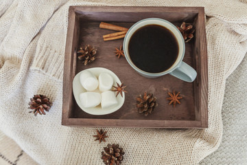 Cup of coffee on rustic wooden tray, sweet marshmallow and warm woolen sweater. Cozy autumn or winter weekend or holidays at home. Fall home decoration with hot drink mug. Hygge morning style concept