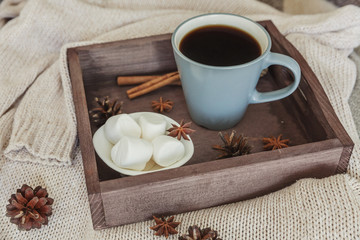 Cup of coffee on rustic wooden tray, sweet marshmallow and warm woolen sweater. Cozy autumn or winter weekend or holidays at home. Fall home decoration with hot drink mug. Hygge morning style concept