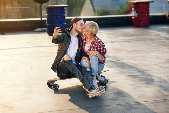 Handsome young couple sitting on longboard skateboard on the rooftop of industrial building and making self photo on smartphone. Modern urban active lifestyle concept.
