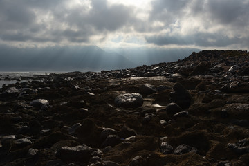 the sun's rays against the backdrop of the mountains and the lava-rocky wet shore in the foreground