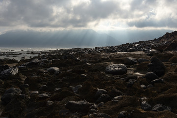 the sun's rays against the backdrop of the mountains and the lava-rocky wet shore in the foreground