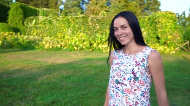 European young woman with long black hair runs away from the camera in slow motion, calling for herself. Attractive girl with dimpled cheeks in a white floral print beckons to follow her. Smiling