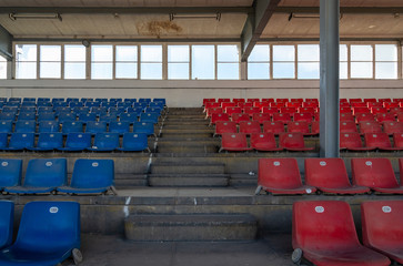 Front view row of blue and red plastic seats on rough concrete different level  floor between stair in old vintage dirty concrete structural stadium with windows frame background without people. 