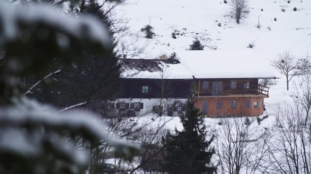 A view of an alpine style house in a forest, covered with snow on a cloudy winter day. A car drives by the house. RACK focus from the house to a close pine branch.