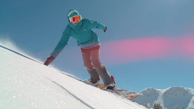 SLOW MOTION, CLOSE UP, LENS FLARE: Fit young Caucasian woman snowboarding off piste in the picturesque sunny Alps does a cool hand drag. Cheerful female tourist carving the freshly fallen powder snow.