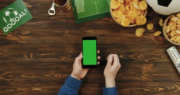 Top view on the black smartphone in male hands being vertically on the wooden desk with football things on it: ball, beer, remote control and chips. Man scrolling and taping on the green screen