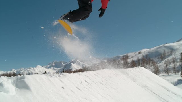 SLOW MOTION, CLOSE UP: Athletic male snowboarding in the spectacular sunny Alps spins in the air while doing a grab. Cinematic shot of pro snowboarder jumping off a kicker and doing a cool trick.