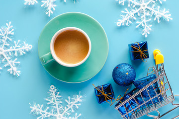 Cup of coffee with Xmas snowflake, gift box, glitter balls and boot ornaments in mini shopping cart or trolley on blue background for drinks and beverage in Christmas day and holidays concept