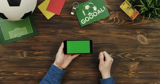 Top view on the black smartphone with chroma key screen being horizontally on the wooden table and male hands scrolling and taping on it. Football ball, goal card and fan ticket lying beside. Green