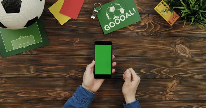 Top view on the black smartphone with chroma key screen being vertically on the wooden table and male hands scrolling and taping on it. Football ball, goal card and fan ticket lying beside. Green