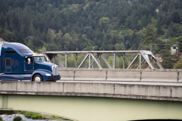 Dark blue big rig bonnet semi truck going on the bridge across the small river