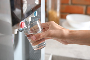 Woman filling glass with water from cooler indoors, closeup