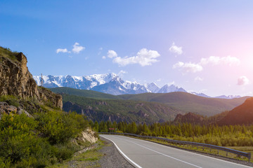 Winding asphalt road near the cliff against the backdrop of high snow-capped mountains with white glaciers on the tops with blue sky and clouds, near the road a fence for security in Altai at sunset