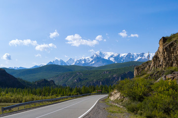 Winding asphalt road near the cliff against the backdrop of high snow-capped mountains with white glaciers on the tops with blue sky and clouds, near the road a fence for security in the Altai