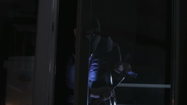 Cunning Caucasian criminal in balaclava picking lock for rob apartment. Man wearing black clothes with gun and flashlight in hands.