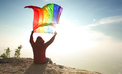 man sitting on mountain top raise rainbow LGBT symbol flag to bright sunny blue sky