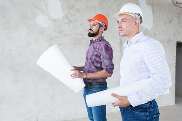 male engineers, architects in helmets stand by the window and hold a roll of paper with a building drawing