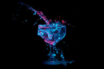 Clear liquid under blue and red lights splashing into a glass goblet isolated on a black background