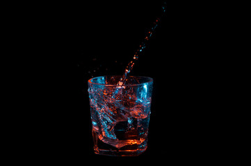 Clear liquid being poured into a spiral in a rocks glass under blue and orange lights isolated on a black background