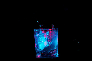 Ice cubes splashing into a rocks glass of liquid under colorful lights isolated on a black background