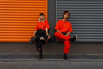 Beautiful fashionable young stylish couple in fashion orange clothes with caps sit near the metal...