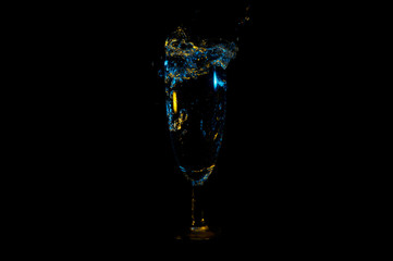 Beverage splashing out of a tall stemmed glass under yellow and blue lights isolated on a black background