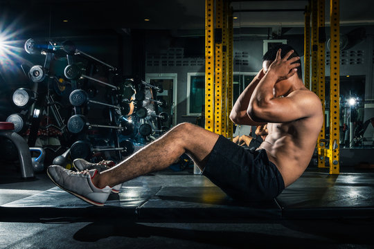Muscular guy doing sit ups at gym with other people in background. Young athlete doing stomach workout in modern gym. Handsome fit man doing crunches at gym.