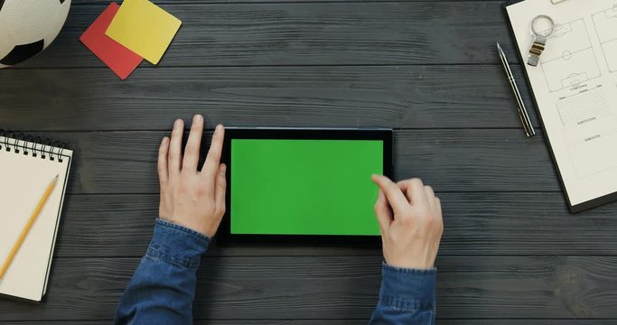 Top view on the black tablet device lying horizontally on the dark grey wooden table of football coach and male hands scrolling and taping on the green screen. Chroma key.
