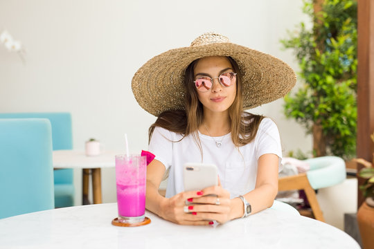 Smiling cute girl in straw hat relaxing in cafe beside glass door holding smartphone in hand.