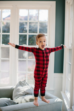 Cute young in pajamas standing on the armrest of a big chair with his hands stretched out