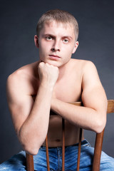 Muscular blond young handsome man sit on chair in blue jeans topless, brow furrowed, fist near face, looks straight,  biceps, sportsman, studio light, low key, black background, close up