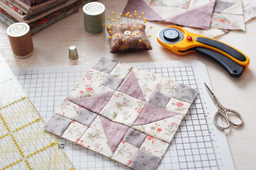 Patchwork block on craft mat, stack of fabric, sewing accessories on white wooden surface