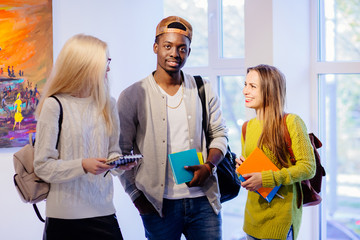 Portrait of three positive, friendly nice look students indoor in college.Hipster black skinned man and two european women spending free time together. Multi ethnic, study abroad, friendship concept.